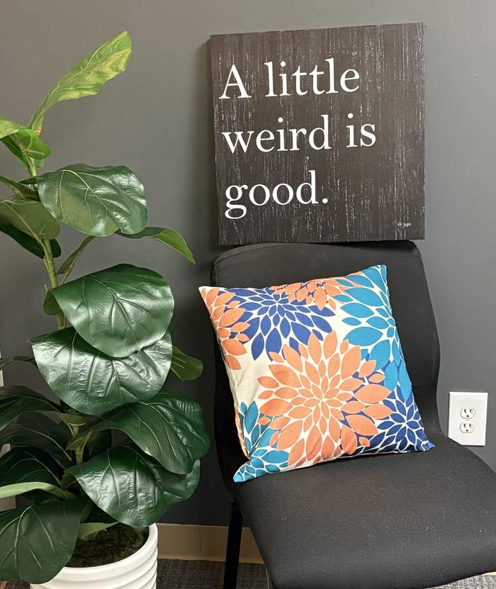 A cozy corner with a plant, a floral pillow, and a sign that reads: A little weird is good