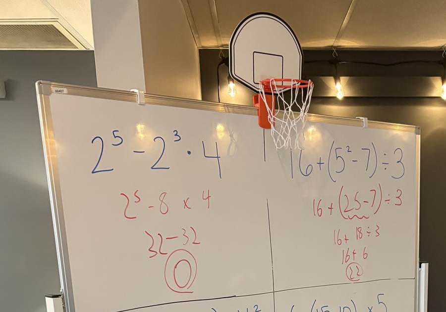A whiteboard of order-of-operations practice problems with a mini basketball hoop above it